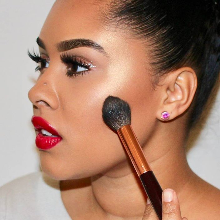 person wearing red lipstick and applying highlighter to cheek with makeup brush