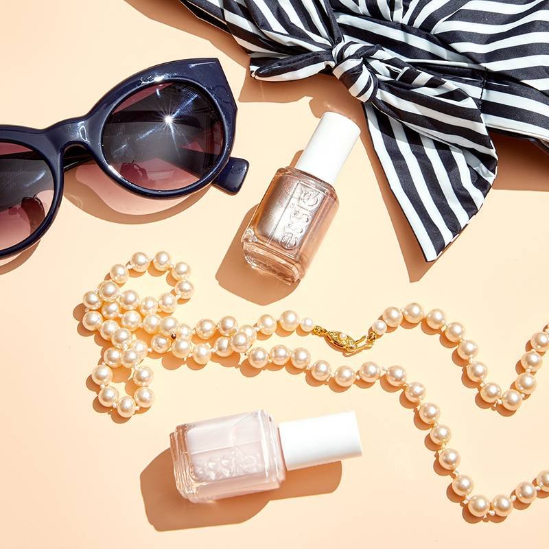 essie nail polish surrounded by sunglasses, a scarf, and and pearl necklace