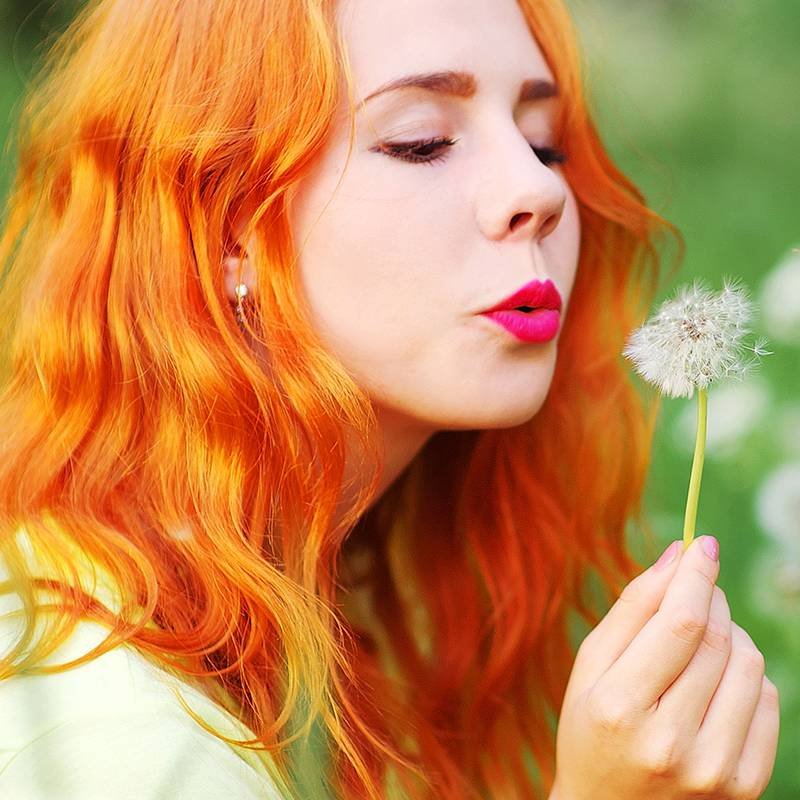 person with red hair wearing pink lipstick and blowing dandelion seed head