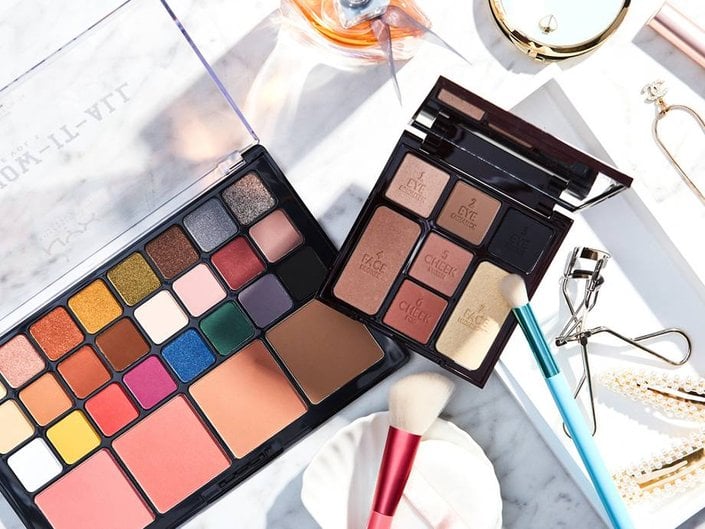 Overhead shot of two makeup palettes on a bathroom counter with makeup brushes, an eyelash curler, hair clips, a necklace and a bottle of perfume
