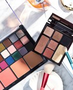 Overhead shot of two makeup palettes on a bathroom counter with makeup brushes, an eyelash curler, hair clips, a necklace and a bottle of perfume