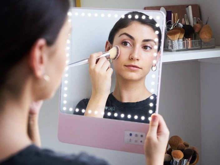 A person holds up a lit-up mirror and applies blush to their cheek with makeup brushes stored in plastic organizers in the background