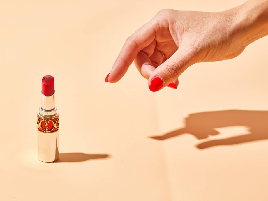 A hand with red nail polish reaching for red YSL Beauté Rouge Volupté Shine Lipstick Balm