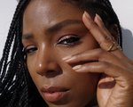 Picture of a person with dark skin wearing nude nail polish, holding their fingers to their face