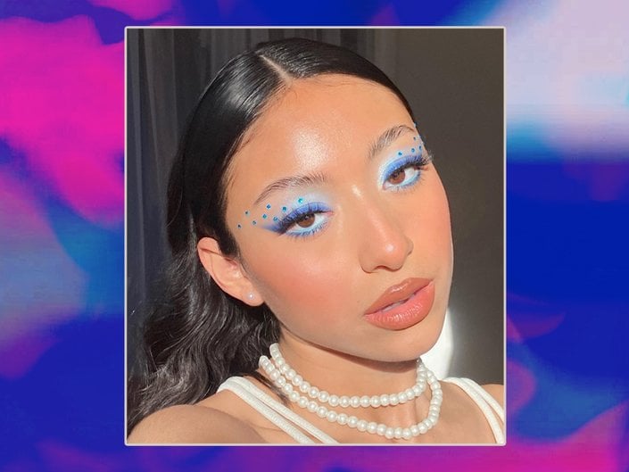Person with black hair wearing blue eyeshadow with blue rhinestones, nude lipstick and a pearl necklace
