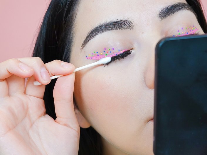 Picture of a person wearing colorful eyeliner and holding a cotton swab to their eyelashes.