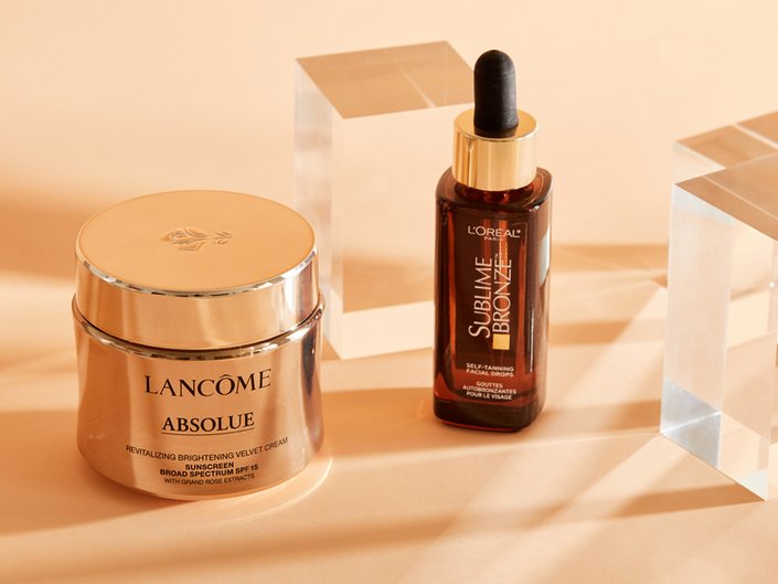 Lancôme Absolu Cream and L’Oréal Paris Facial Tanning Drops photographed on a peach background. 