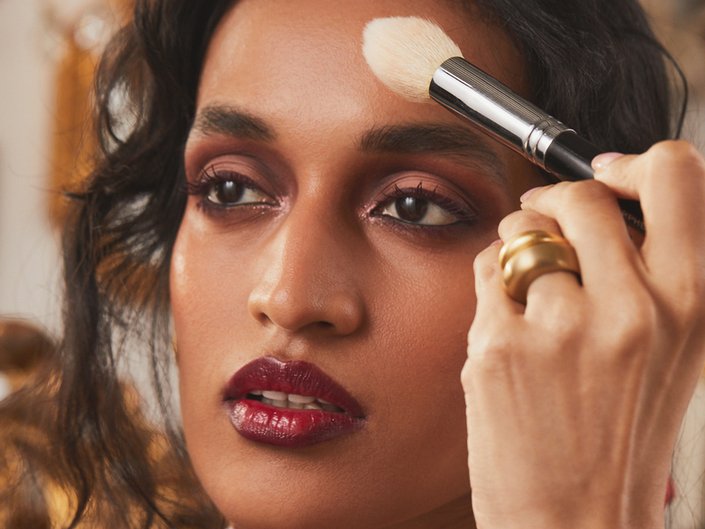 person holding foundation brush to forehead and wearing burgundy eyeshadow and lipstick