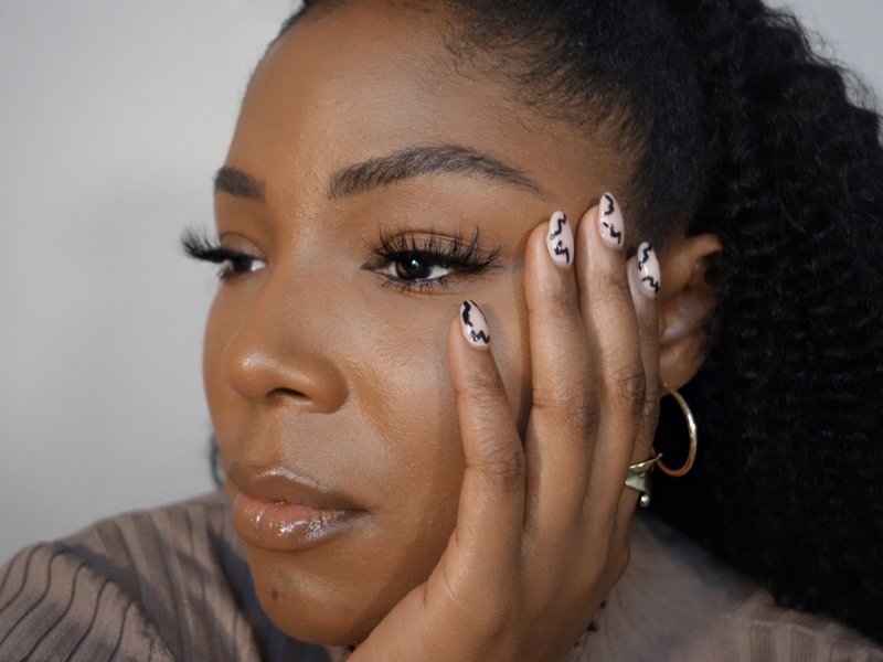 Person wearing false eyelashes looking to the side of the camera with their face resting in their hand and their nails painted with a black design
