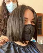 Selfie of person posing with their hairstylist with half of their dark hair cut and the other half in a pigtail. Both people are wearing masks and they are in a salon setting.
