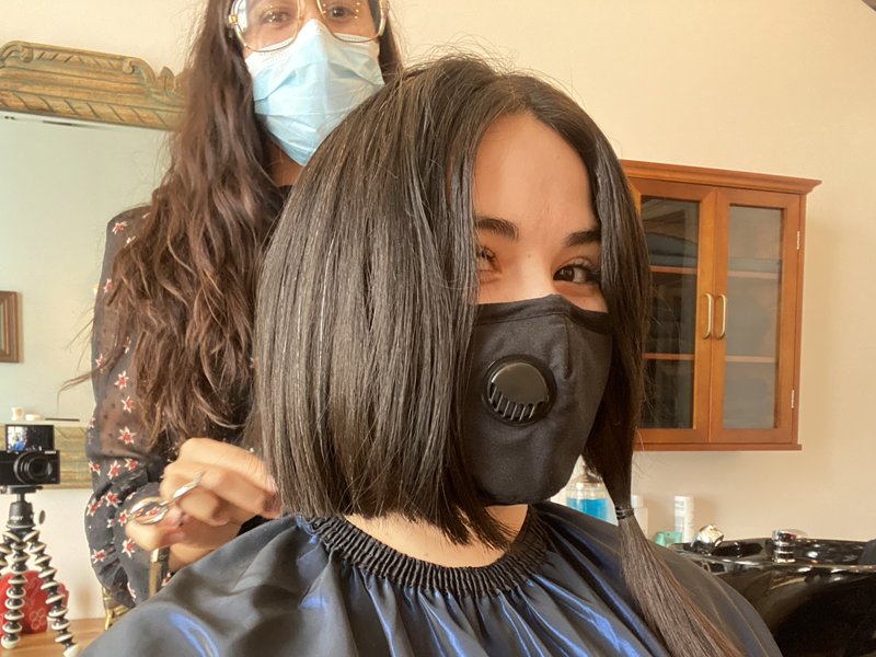 Selfie of person posing with their hairstylist with half of their dark hair cut and the other half in a pigtail. Both people are wearing masks and they are in a salon setting.