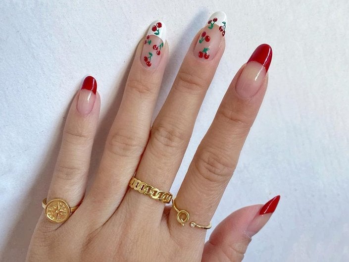 A picture of a hand with a red-and-white French tip and cherry-printed manicure against a white wall