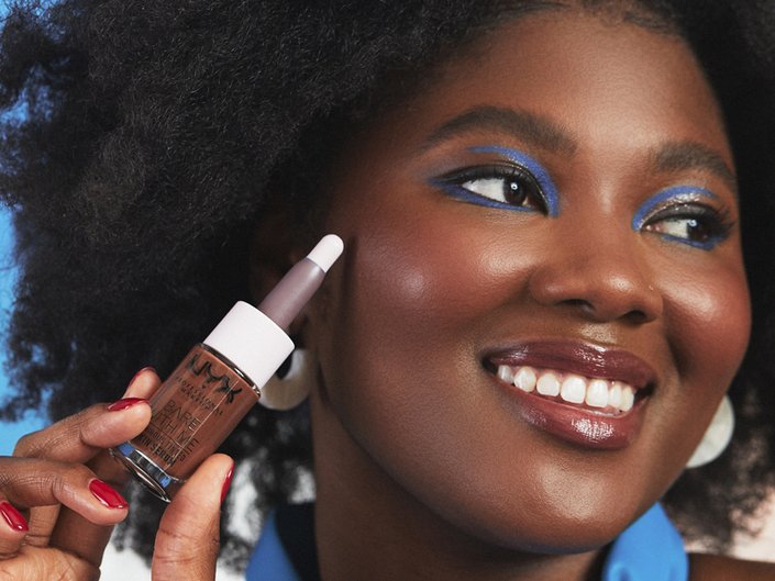 person wearing blue graphic eyeshadow and holding NYX Professional Makeup Bare With Me Luminous Tinted Skin Serum