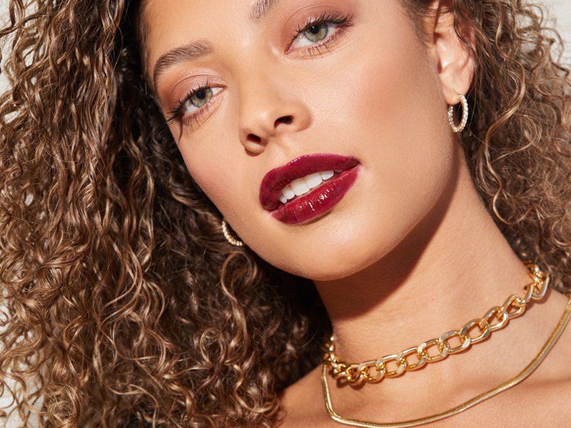 A close-up photo of a person with curly hair wearing red lip gloss and gold jewelry.