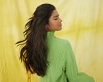 Person with long dark hair wearing white floating eyeliner and a green turtleneck sitting in a chair against a yellow background