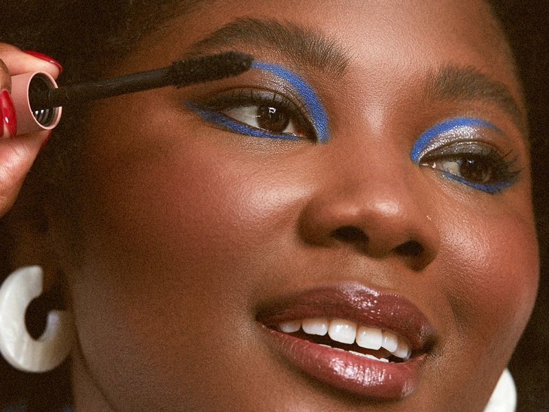 person wearing blue graphic eye makeup holding mascara wand up to eyelashes