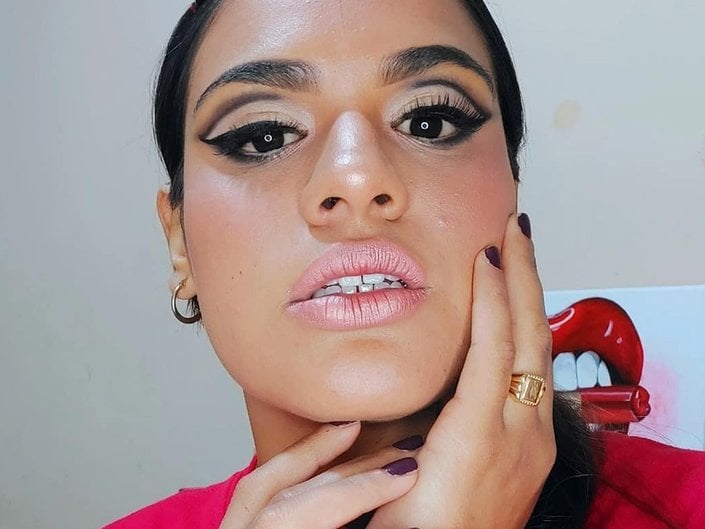 A person with dark hair pulled back looking at the camera, wearing a full face of makeup and cut crease eyeshadow