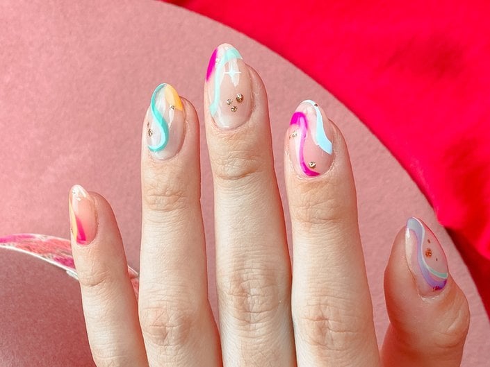 A close-up of a hand with nails painted in swirls of blue, purple, pink and yellow polish, accented with nail gems.