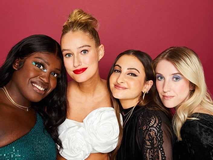 Four Makeup.com Beauty Editors Posing Together against a maroon red background, each styled with different makeup looks.