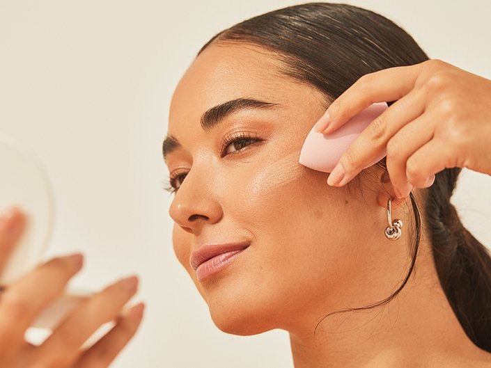 Person looks in a compact mirror and applies foundation to their cheekbone with a makeup sponge