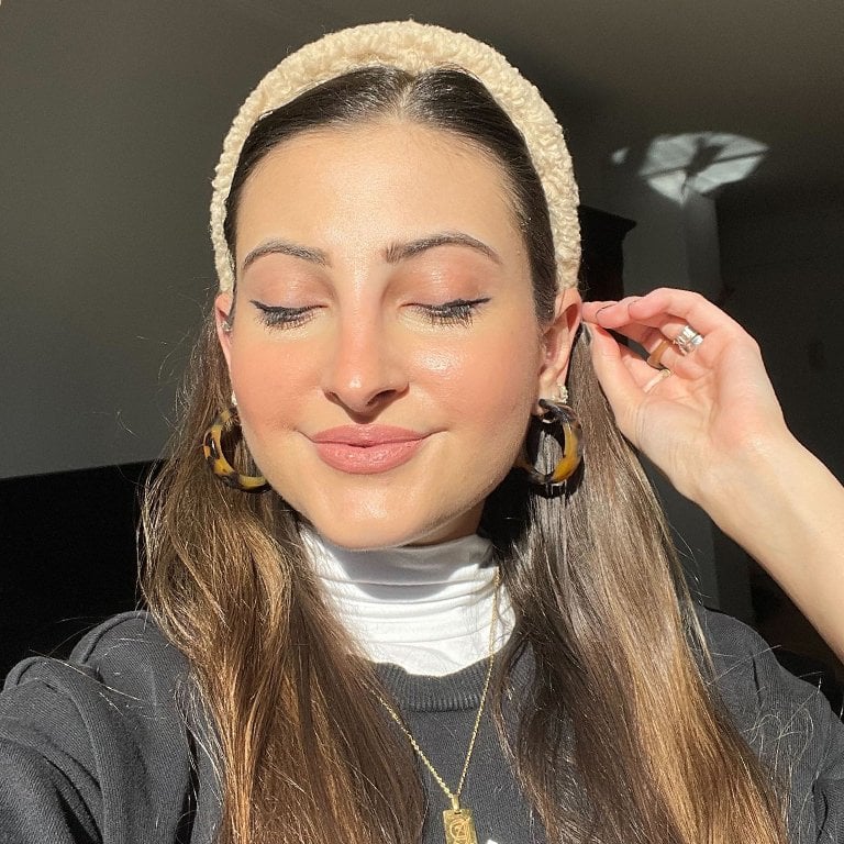 Selfie of Makeup.com editor with eyes closed and hand to ear wearing a headband and dewy makeup with blush and pink eeyshadow