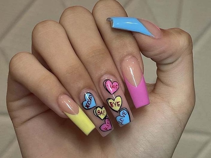 Hand with long rectangular nails with yellow, pink and blue French tips and decorated with colored candy hearts