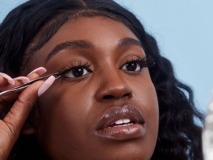 A person looking to the side of the camera applying false eyelashes to their lash line with tweezers