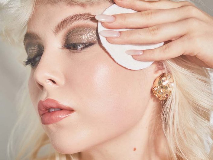 young person with blonde hair holding up a cotton pad to glittery eye makeup