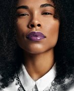 Person with black curly hair looking at the camera and wearing shiny purple lipstick. 