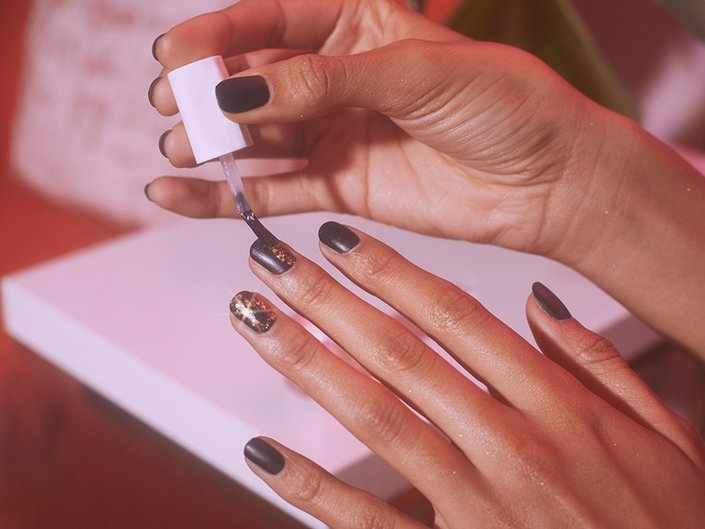 Photo of a person’s hands. Their fingernails are painted black, and they are applying a top coat of glittery nail polish