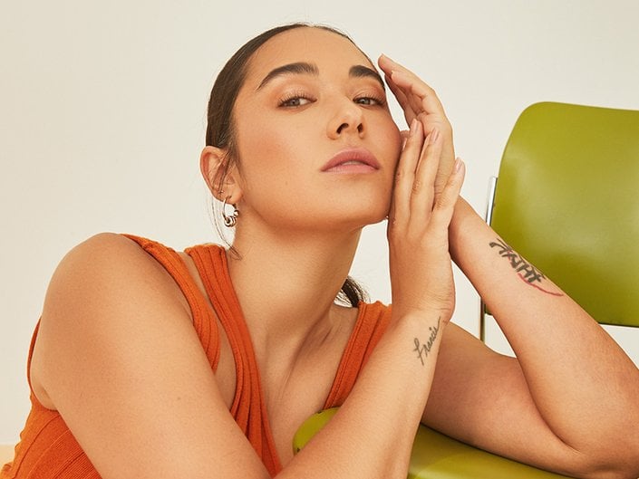 A person in an orange shirt leaning on a green chair with their hands touching their face