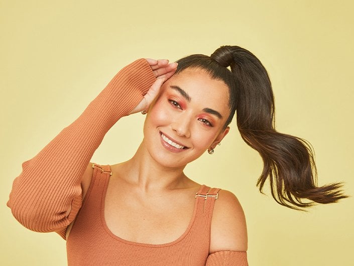 Person smiling and wearing an orange off-the-shoulder sweater with their dark hair in a high ponytail