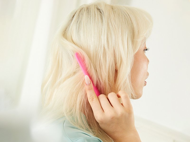 Person faces away from the camera and brushes a pink comb through bleached, platinum blonde hair
