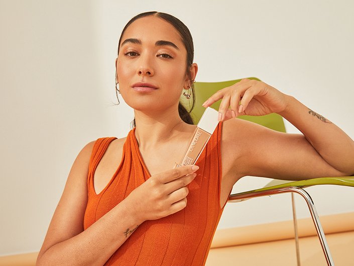 Photo of person in an orange shirt with their dark hair pulled into a slicked back ponytail. They are leaning against a green chair and holding the Lancôme Teint Idole Ultra Wear Care and Glow Foundation.