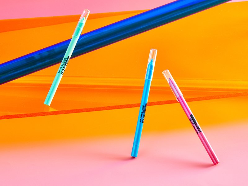 Photo of the Maybelline New York Tattoo Studio Long-Lasting Eyeliner Pencils in bright green, blue and pink on a bright orange background