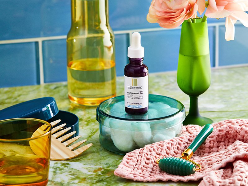 bathroom counter with La Roche-Posay 10% Pure Niacinamide Serum, a jar of cotton balls, a jade face roller, a comb and more