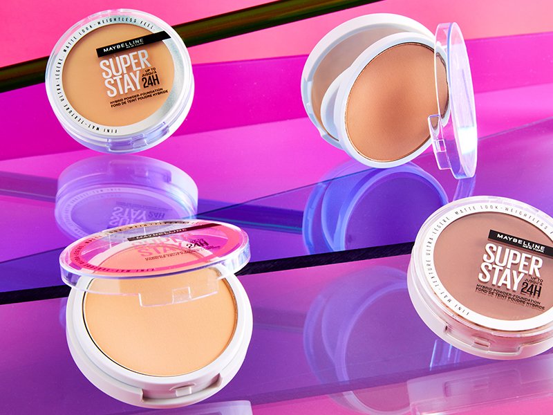 four compacts of the maybelline super stay powder foundations laid out on a neon purple and pink background