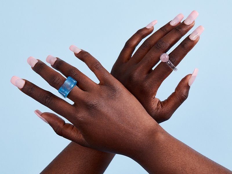 Two hands with rings and long pink square-shaped nails, crossed over each other against a blue background