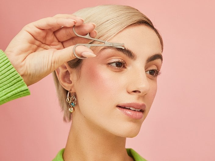 Person with blonde pixie cut and brunette eyebrows looking off to the side of the camera trimming their eyebrows with scissors.