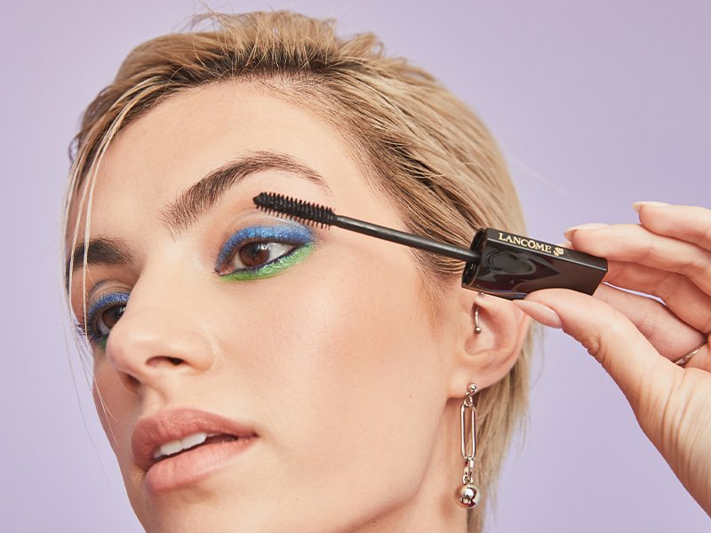 person with blonde pixie cut and blue eyeshadow holding up Lancôme Le 8 Hypnôse Mascara application blush up to eyelashes
