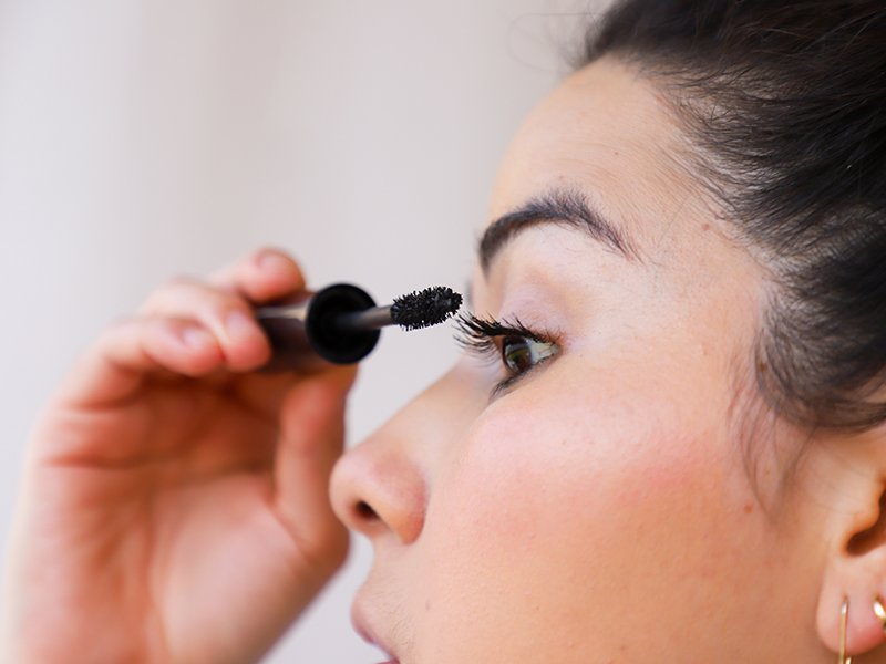 Close-up of person with dark hair in profile applying mascara to their eyelashes 