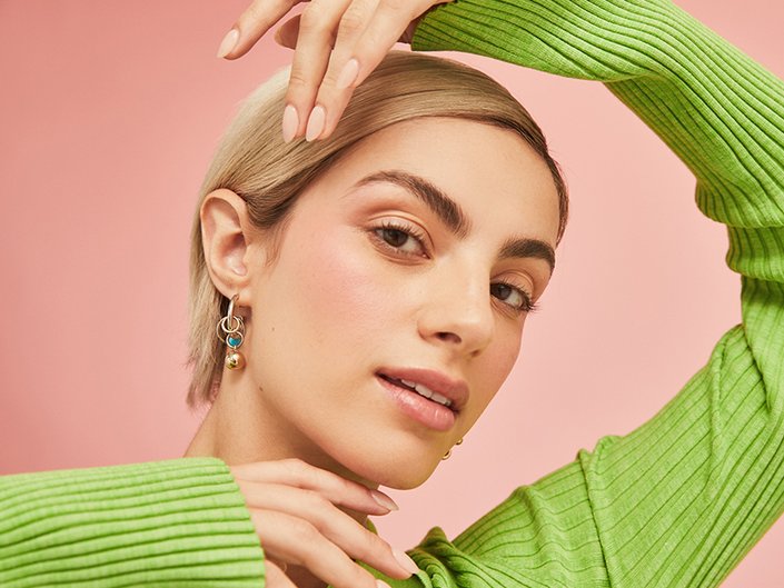Person with blonde pixie cut wearing a green sweater holds their hand over their head near their eyebrows