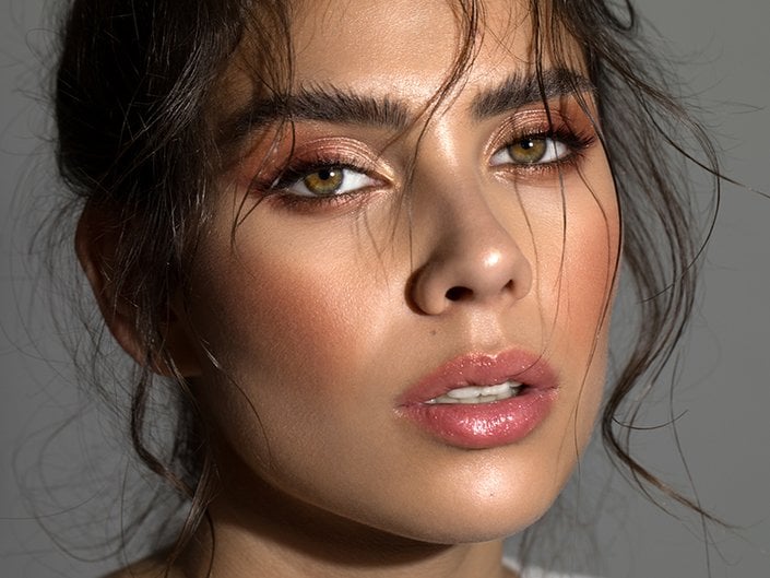 Close up portrait of a person with green eyes, strands of dark hair in their face, shimmery eyeshadow and pink lipstick