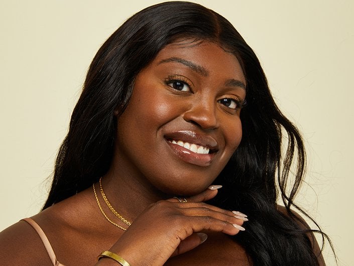 Photo of a person with long dark hair and dewy, clean makeup smiling at the camera and holding their hand under their chin