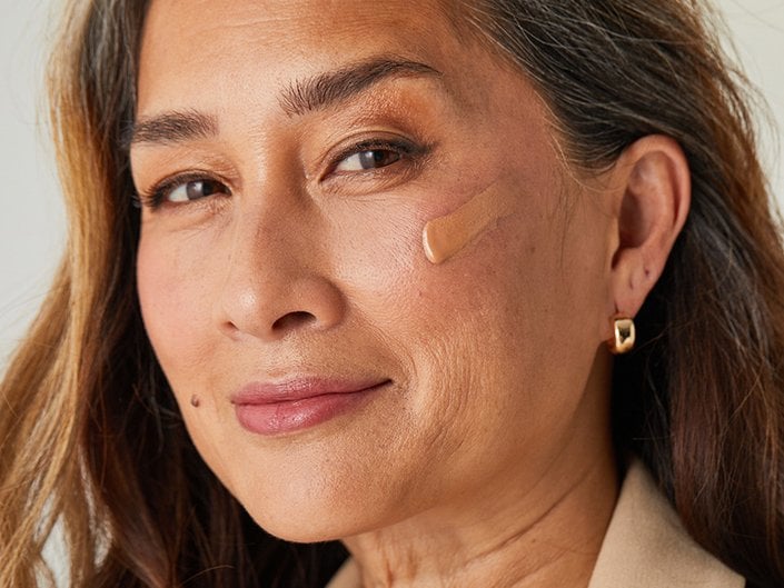 Person with long brown hair looking at the camera with foundation makeup swatched on their cheek