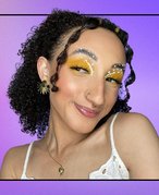 A photo of a person with brunette curly hair wearing electric yellow eyeshadow with pearl appliques. 