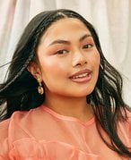 person with long dark hair with two face-framing braids and peach-colored makeup matching a peach-colored shirt