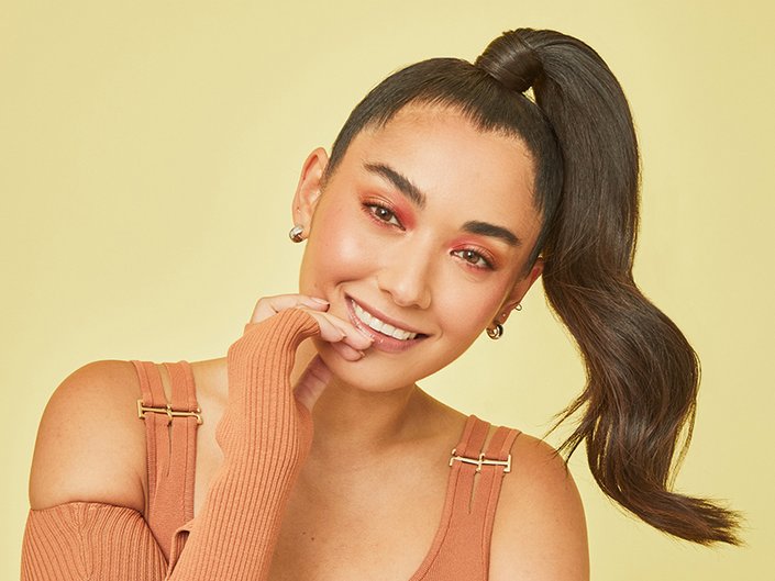 Person with long dark hair in a slicked back high ponytail, smiling at the camera