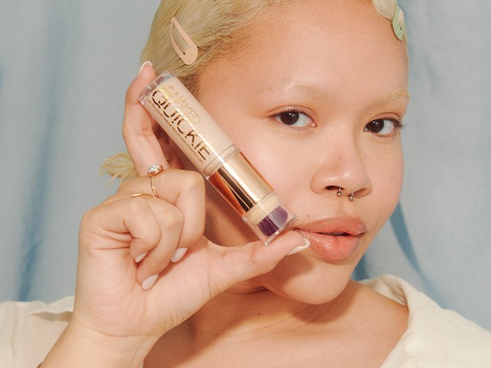 Model with bleached hair and eyebrows looking at the camera and holding up Urban Decay Quickie Concealer