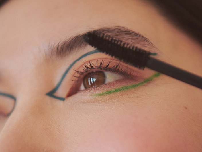 An upclose image of someone applying mascara to their lashes. The person is wearing blue and green floating eyeliner.